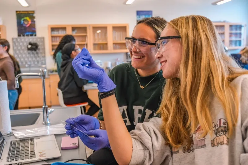 Two students in lab goggles laugh at the bench