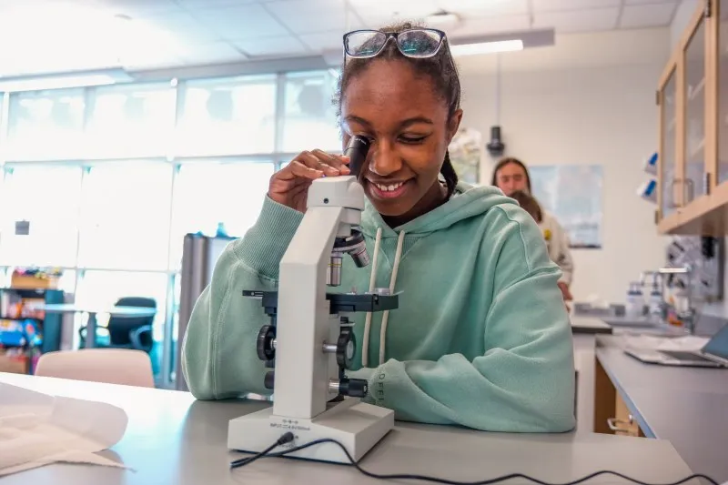 A student smiles as she looks into a microscope in a science lab